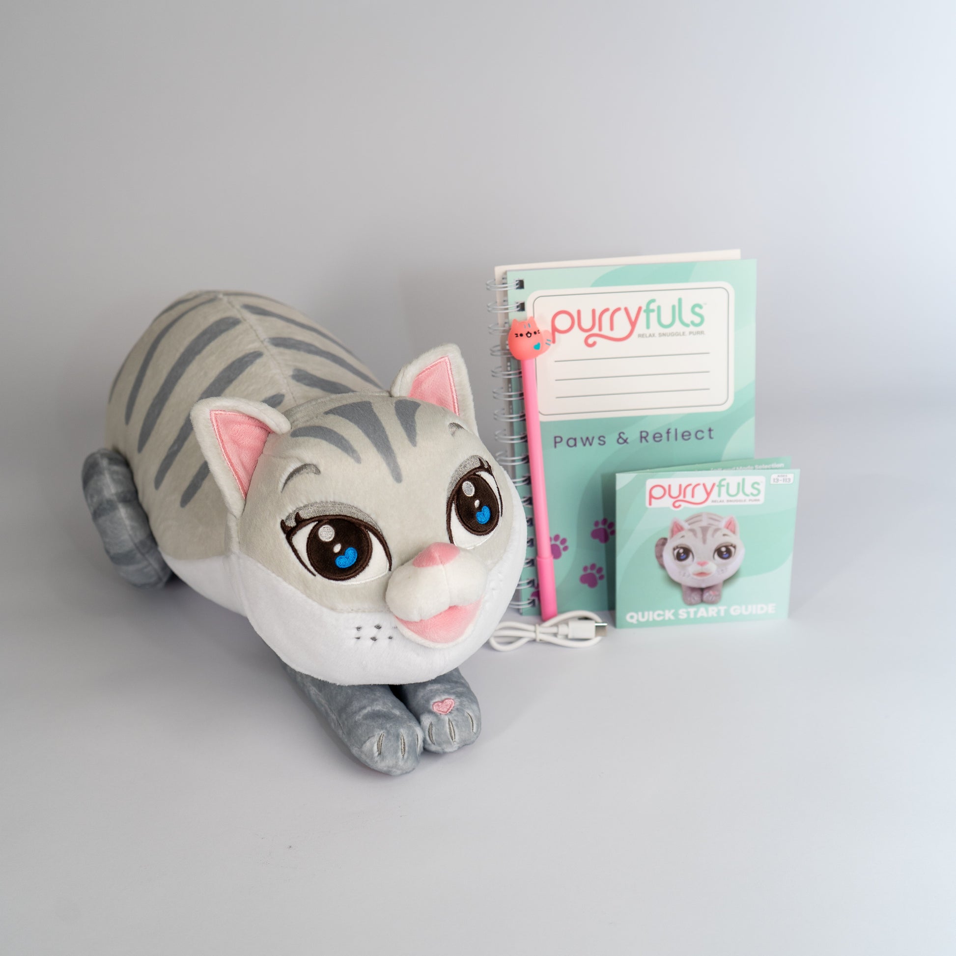 A plush toy cat in a lying-down pose with Siamese tabby coloring, large blue eyes, pink inner ears, and a pink nose, shown beside a mint-green 'purrfuls Paws & Reflect' notebook, a matching pen, charging cord, and instruction booklet on a light gray background.