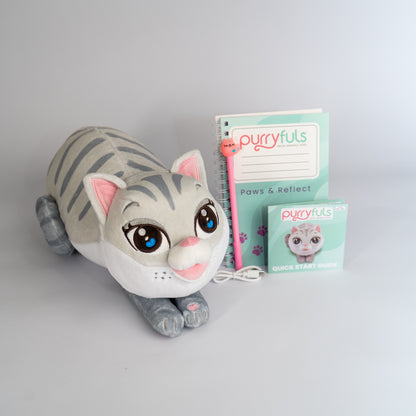 A plush toy cat in a lying-down pose with Siamese tabby coloring, large blue eyes, pink inner ears, and a pink nose, shown beside a mint-green 'purrfuls Paws & Reflect' notebook, a matching pen, charging cord, and instruction booklet on a light gray background.